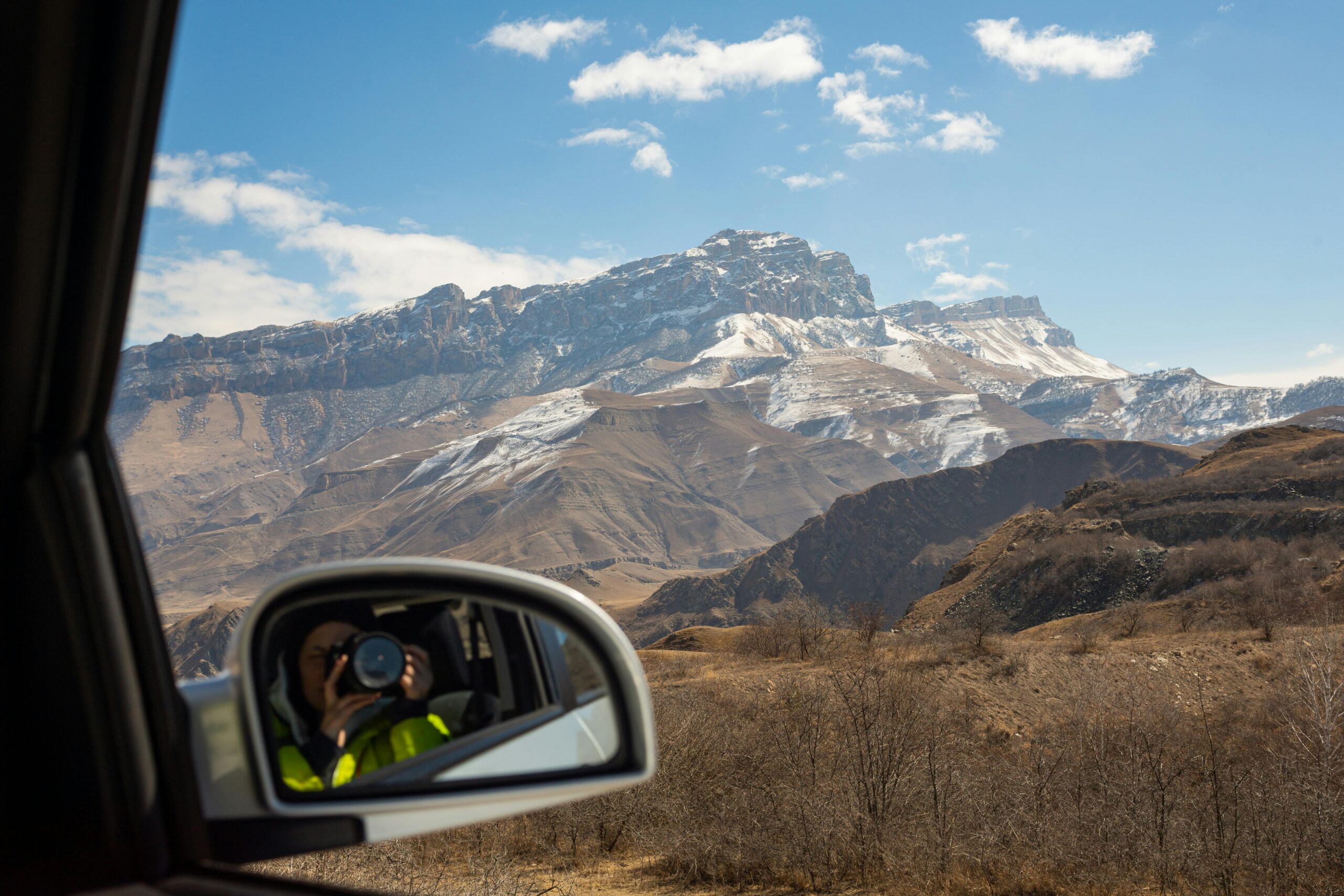 A photographer captures snow-capped mountains through a car's side mirror, blending technology with nature.